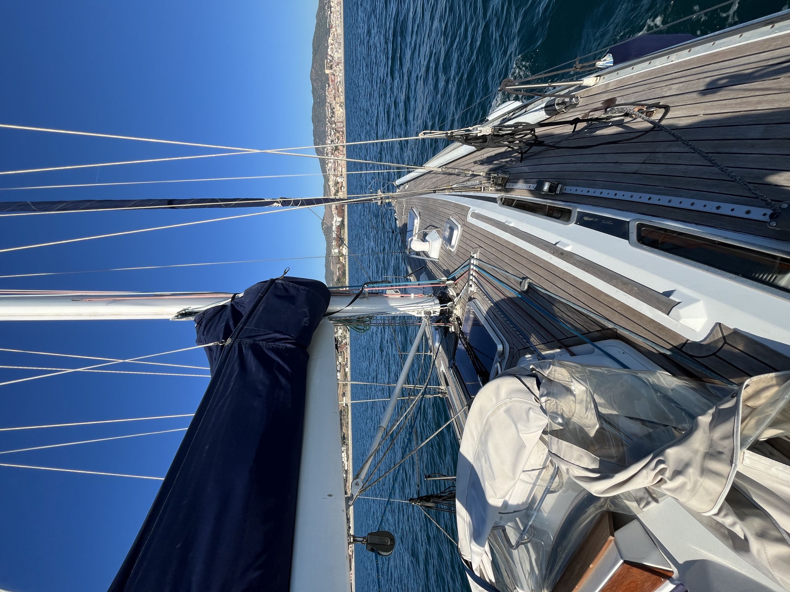 A sailboat's deck with multiple masts and rigging, a cabin with windows, and a view of the coastline.