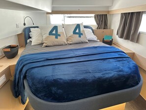 Una cama con cubierta azul, dos almohadas con el número 44, una mesita de noche y una ventana con cortinas.