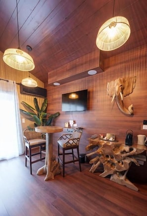 Un intĂ©rieur en bois avec une table ronde, des chaises, une sculpture reprĂ©sentant la tĂȘte dâun Ă©lĂ©phant fixĂ©e au mur, et des luminaires en fibres tressĂ©es.