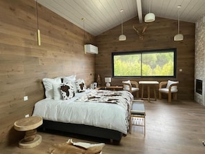 A wooden interior with a bed, a table, chairs, and a large window.