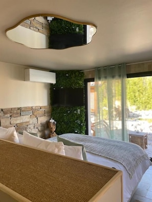 Une chambre avec un lit, un miroir fixé au mur, un mur en pierre, une petite statue et une vue sur la verdure à l’extérieur.