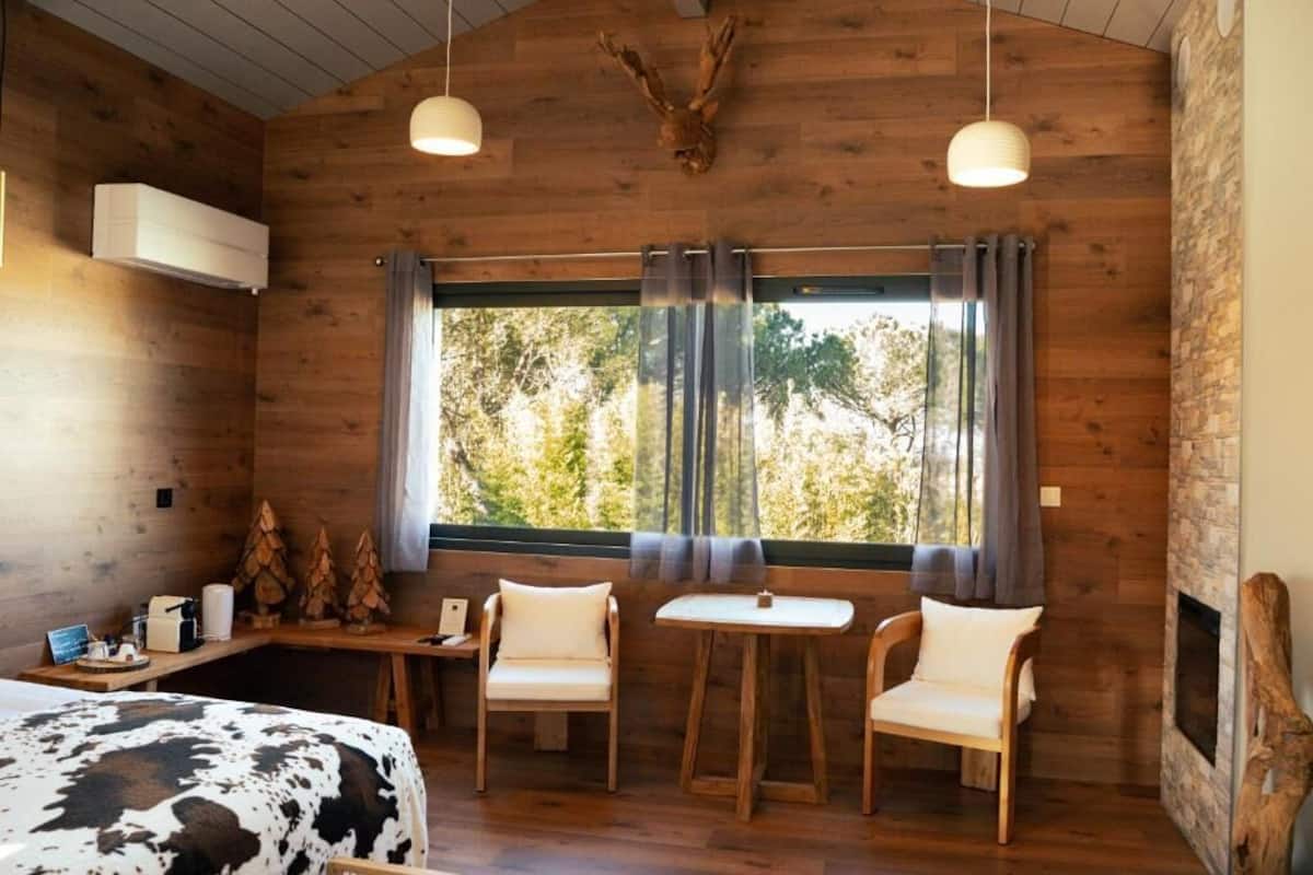 A wooden cabin room with a bed, two chairs, a small table, a window with curtains, and a mounted deer head.