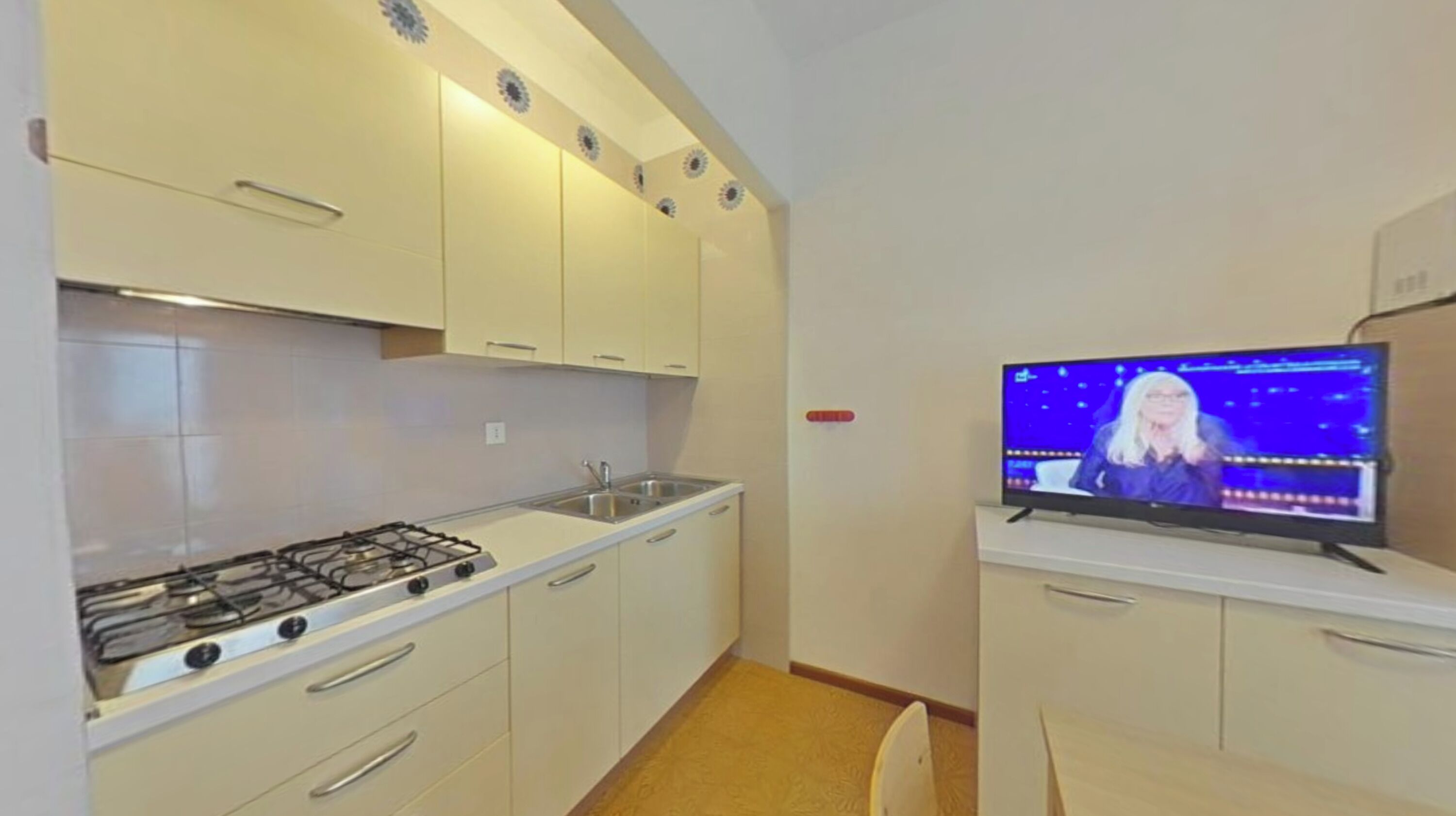 Une petite cuisine avec des meubles blancs, une cuisinière à gaz et un téléviseur diffusant un programme d’information.