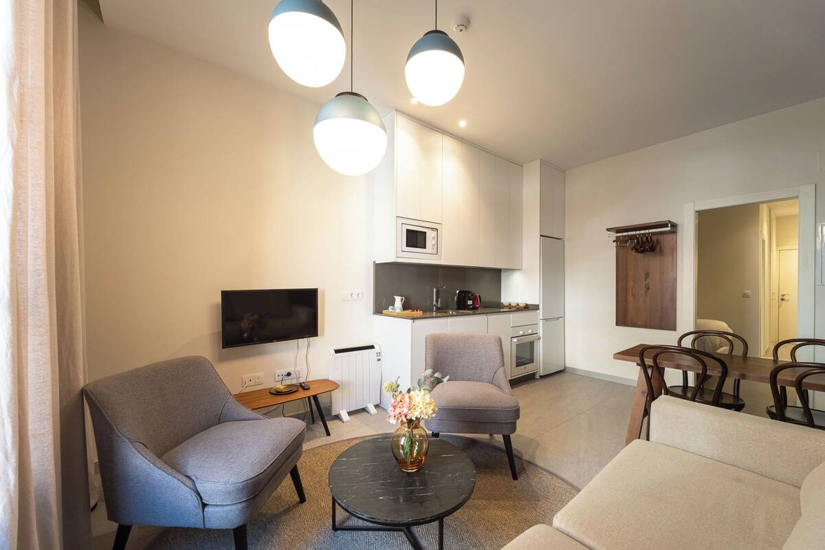 A modern living room with a flat-screen TV, two grey armchairs, a round coffee table with a vase of flowers, and a dining area with a wooden table and chairs.