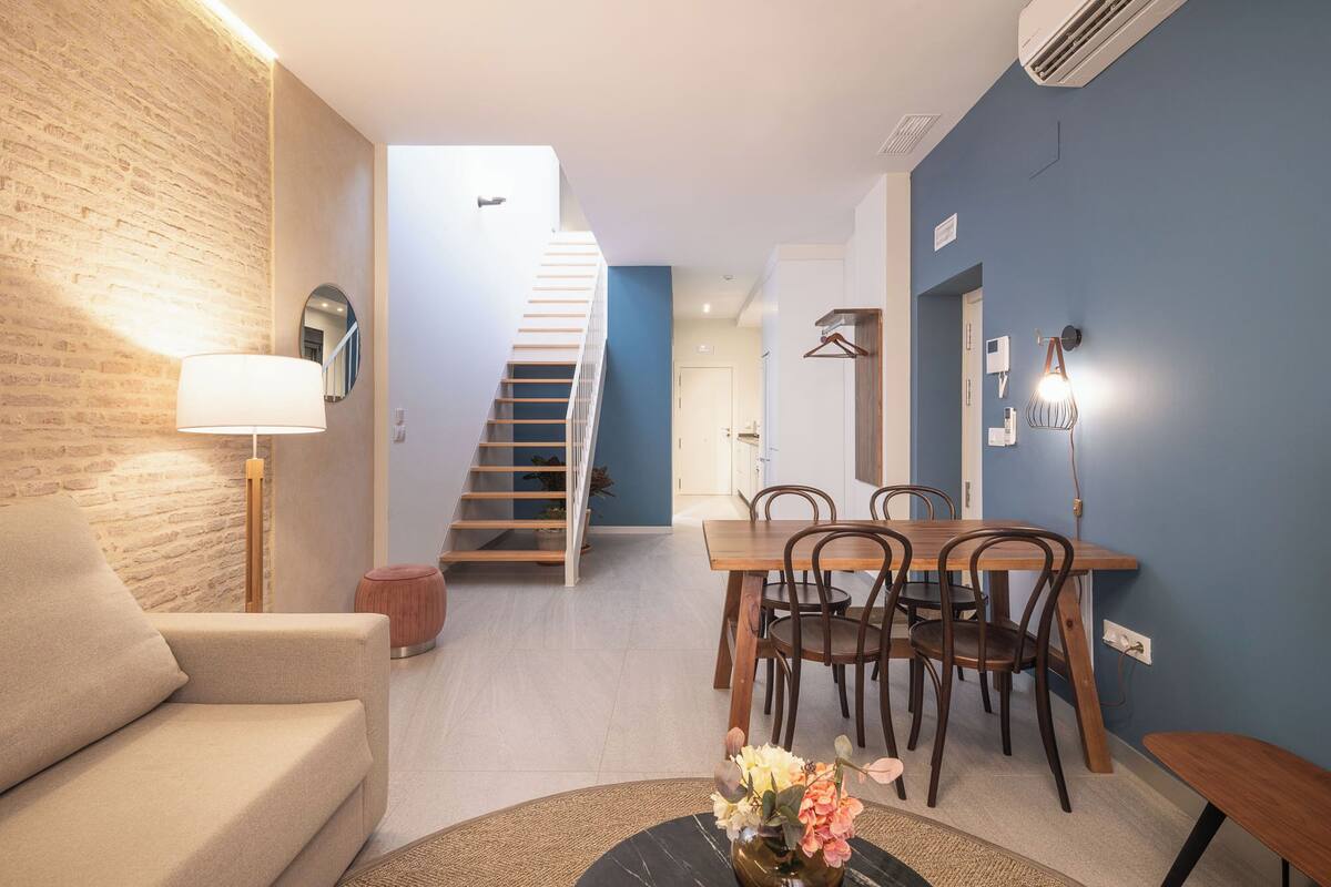 A modern living room with a sofa, a dining table with chairs, a staircase, and a hanging light fixture.