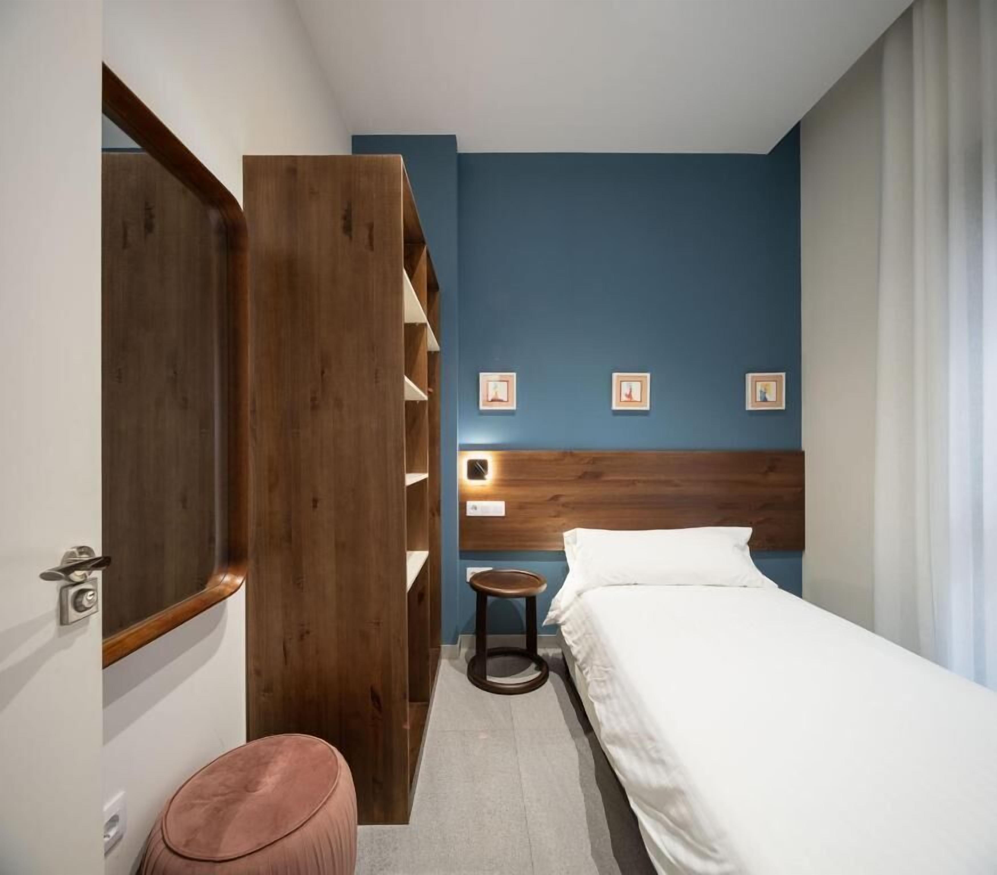 Ein Hotelzimmer mit einem Bett, einem Holzschrank, einem kleinen Hocker und einem Nachttisch mit Lampe.