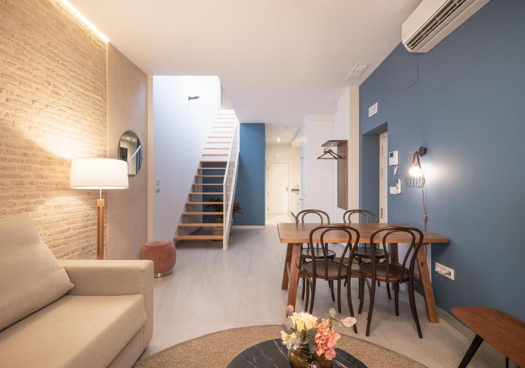 Una sala de estar con sofá, una mesa de comedor con sillas, una escalera y un aire acondicionado instalado en la pared.