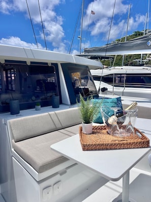 A yacht's interior with a cushioned seating area, a small potted plant, a woven tray with glassware, and a view of other yachts.