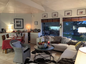 A spacious living area with a dining table, wicker chairs, a sofa with patterned cushions, a round coffee table, and framed pictures on the wall.