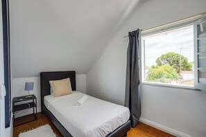 Ein Schlafzimmer mit einem Bett, einem Nachttisch, einer Lampe, einem Fenster mit Vorhängen und Blick auf Bäume und Gebäude.