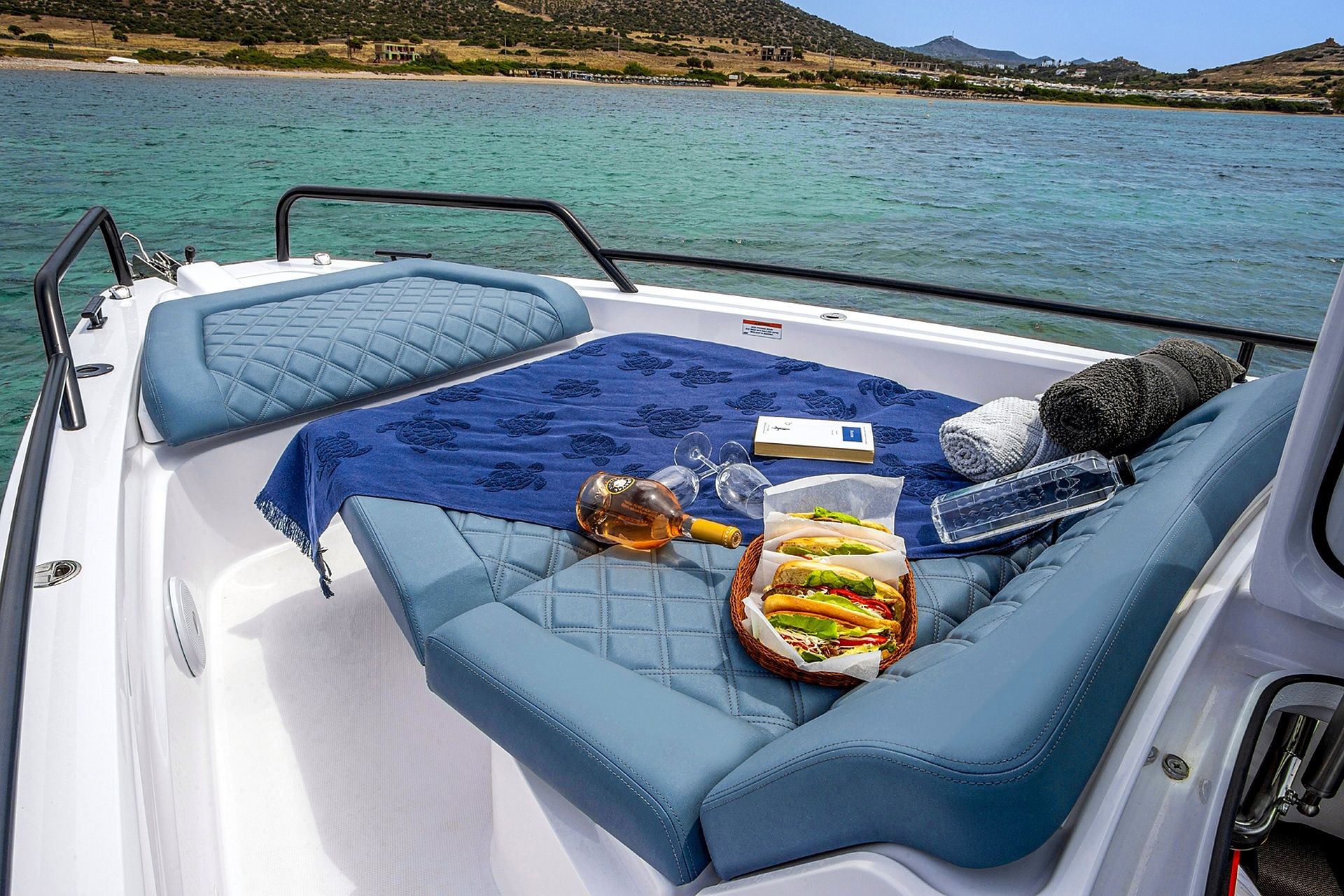 A boat deck with a cushioned seating area, a blanket, a bottle, glasses, and a basket with food.