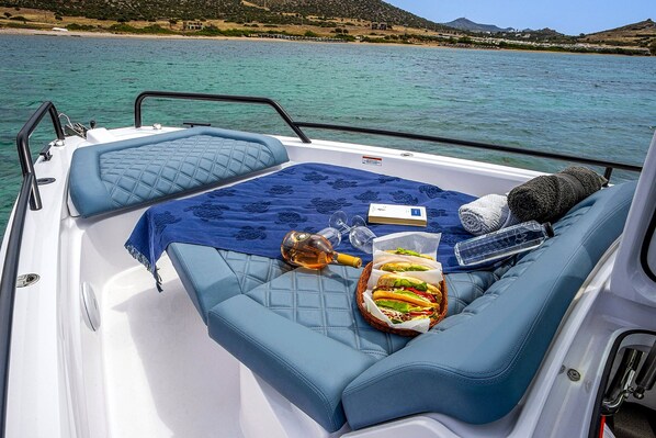Een scheepdek met een zachte zithoek, een plaid, een fles, glazen en een mand met eten.