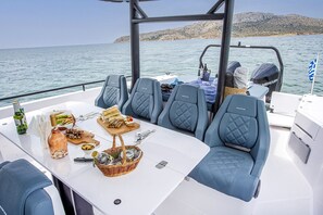 A boat deck with seating, a table set with food and drinks, and a view of the sea and hills. - Sun-Kissed Sailing Adventure in the Stunning Islas Cícladas (Paros)
