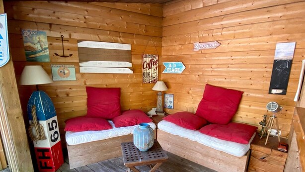 Interior acogedor de una cabaña con piso de madera, dos bancos tapizados en rojo, una mesita con un jarrón azul y varios letreros en las paredes.