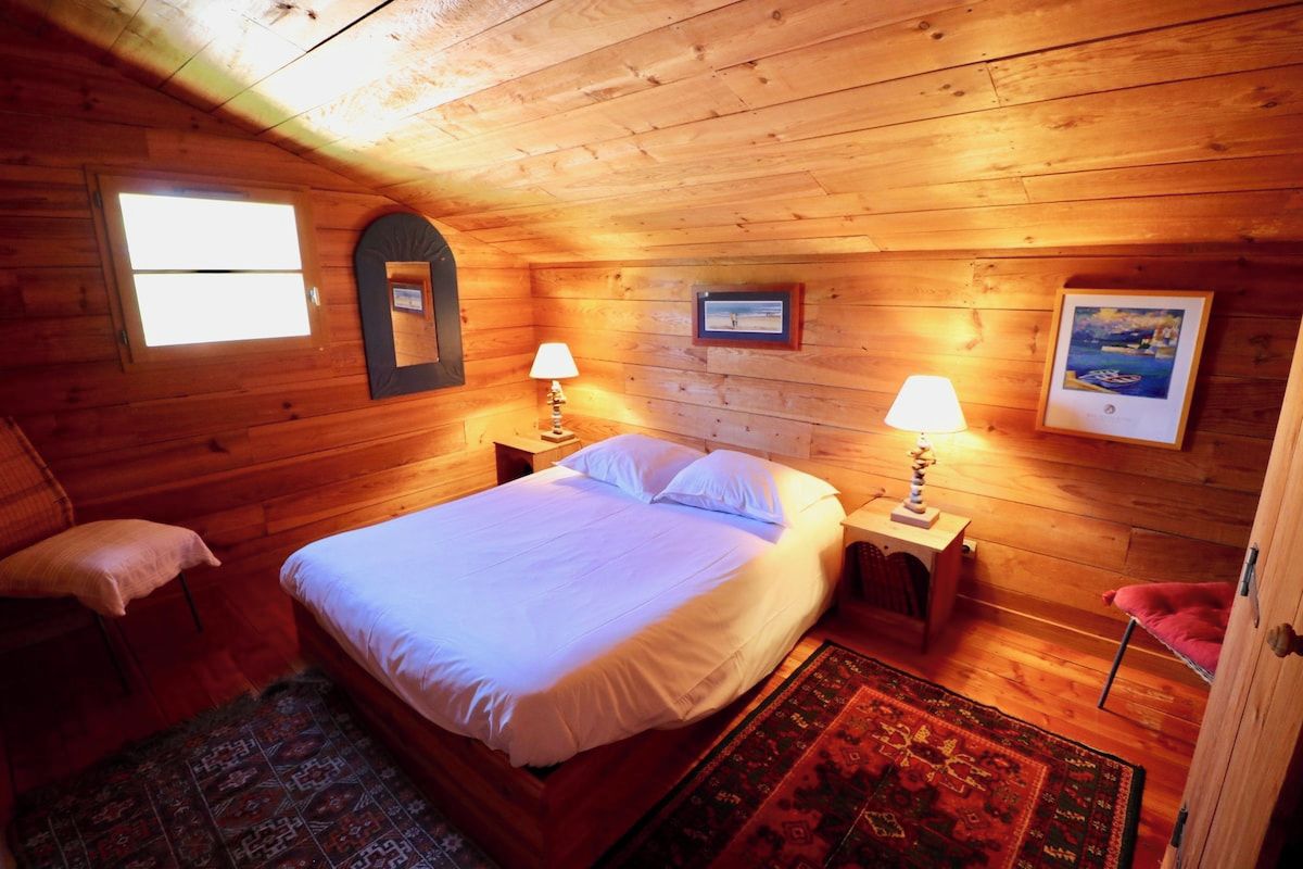 A cozy wooden cabin bedroom with a bed, two bedside tables, a nightstand, a window, and a door.