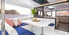 Un intérieur de bateau moderne comprenant un coin repas, des sièges avec des coussins, une table avec un plateau de fruits et une vue sur l’eau.