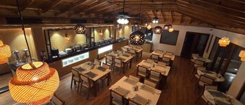 Un restaurante con mesas y sillas de madera, iluminación colgante y una barra con productos alimenticios.