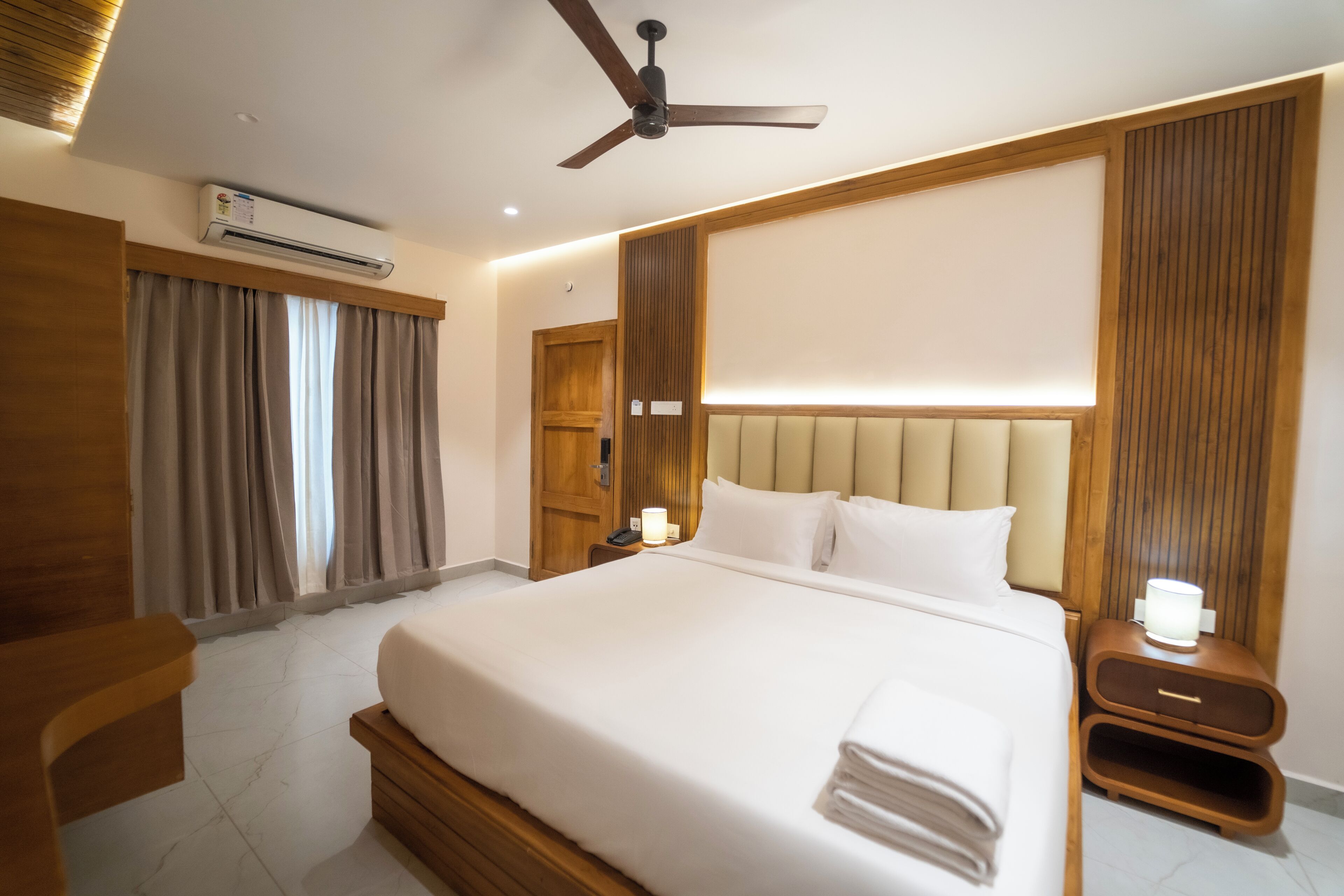 A hotel room with a large bed, a ceiling fan, an air conditioning unit, a bedside table with a lamp, and a wooden headboard.