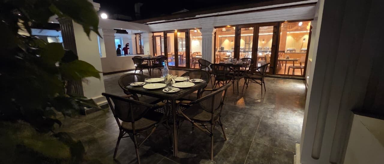 Un espace de restauration en plein air avec des tables et des chaises, une cuisine visible à travers des portes vitrées et une plante au premier plan.