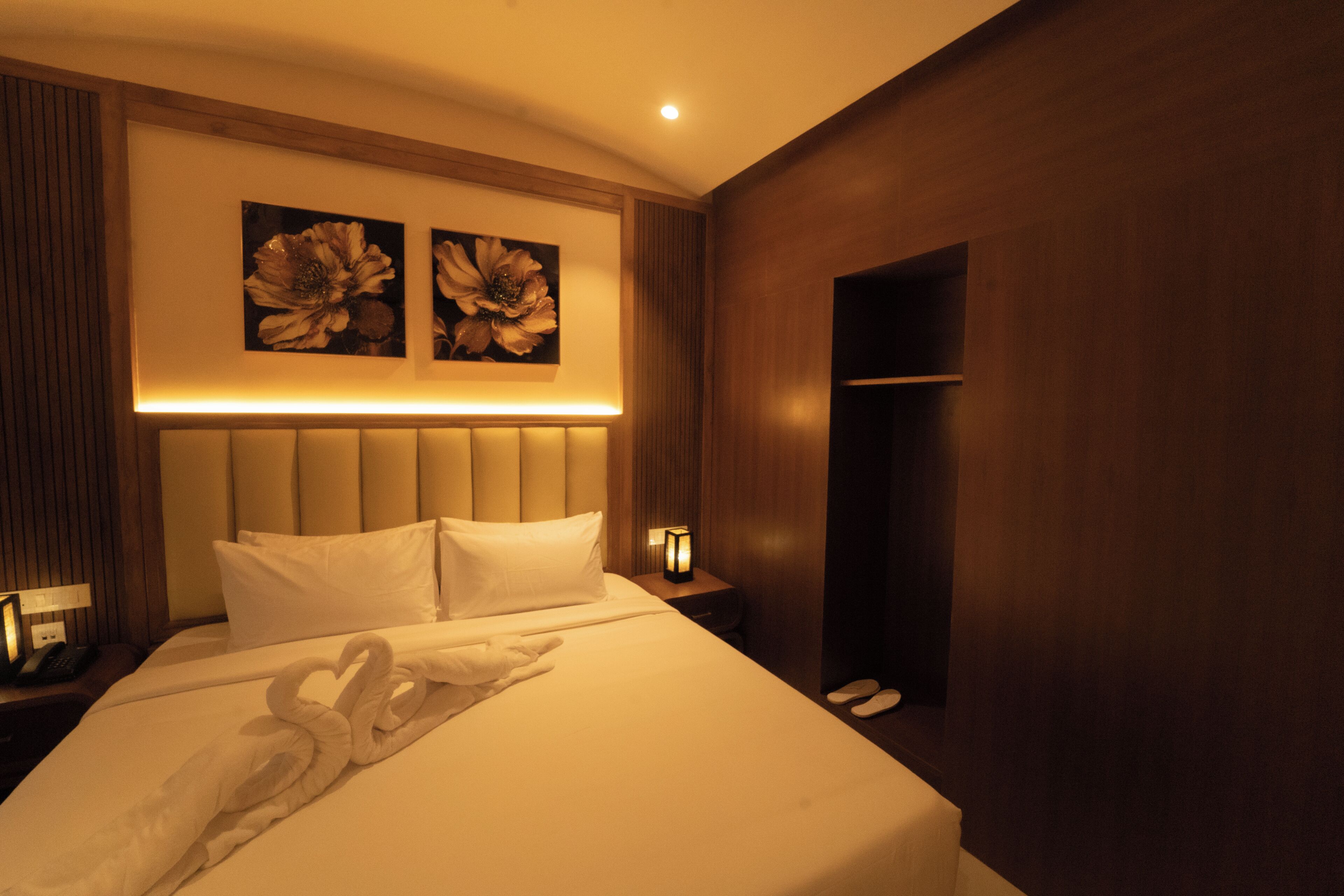 A hotel room with a large bed, a headboard, two wall-mounted lamps, and two floral paintings above the bed.