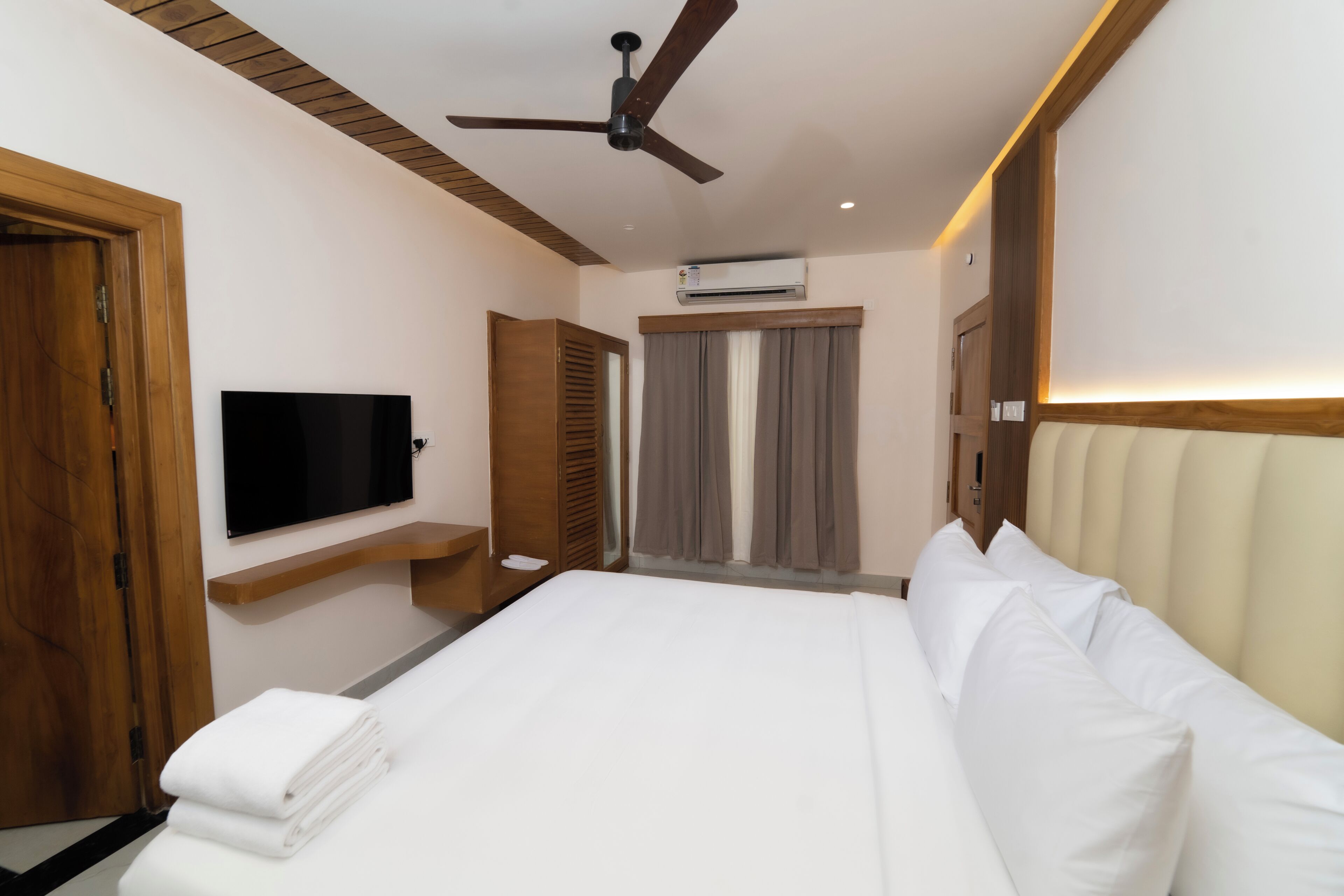 A hotel room with a bed, a ceiling fan, an air conditioning unit, a TV, and wooden doors.