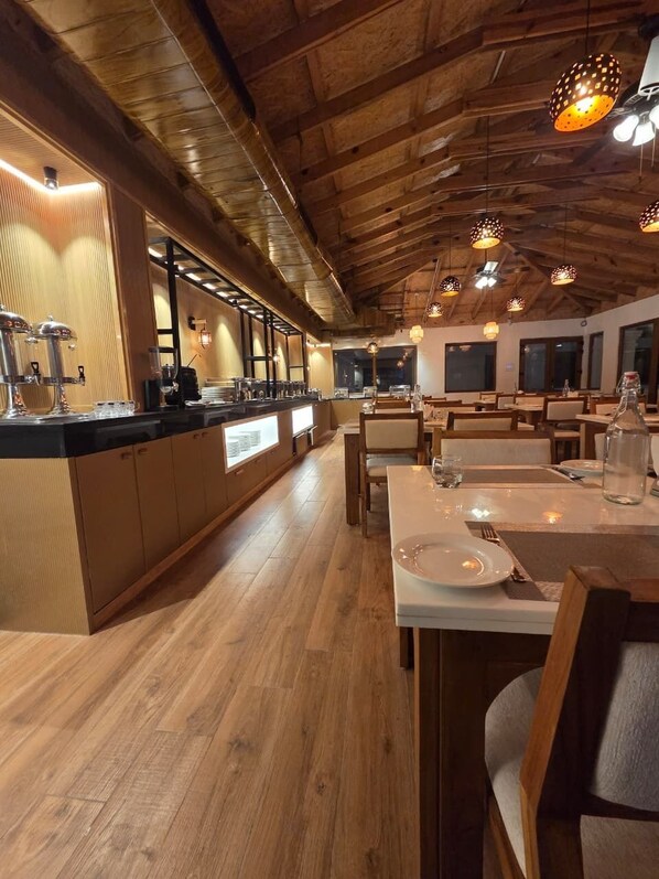 A restaurant with wooden ceilings, pendant lighting, a long counter with a coffee machine, and tables set for dining.