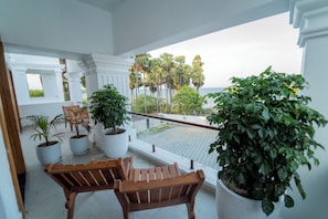 A balcony with potted plants, wooden chairs, and a view of palm trees and the sea.