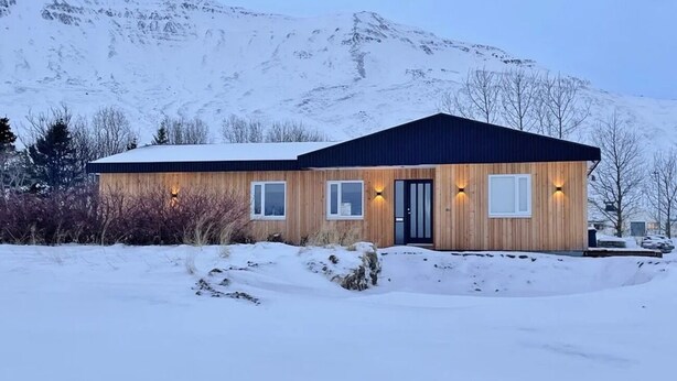 Una cabaña de madera con techo oscuro, ventanas e iluminación exterior, con una imponente montaña nevada al fondo.