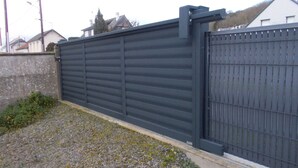 A large, dark-colored metal gate with a solid wall and a metal fence on top. - Charming, cosy villa 5 minutes' walk from the beach (Jullouville)
