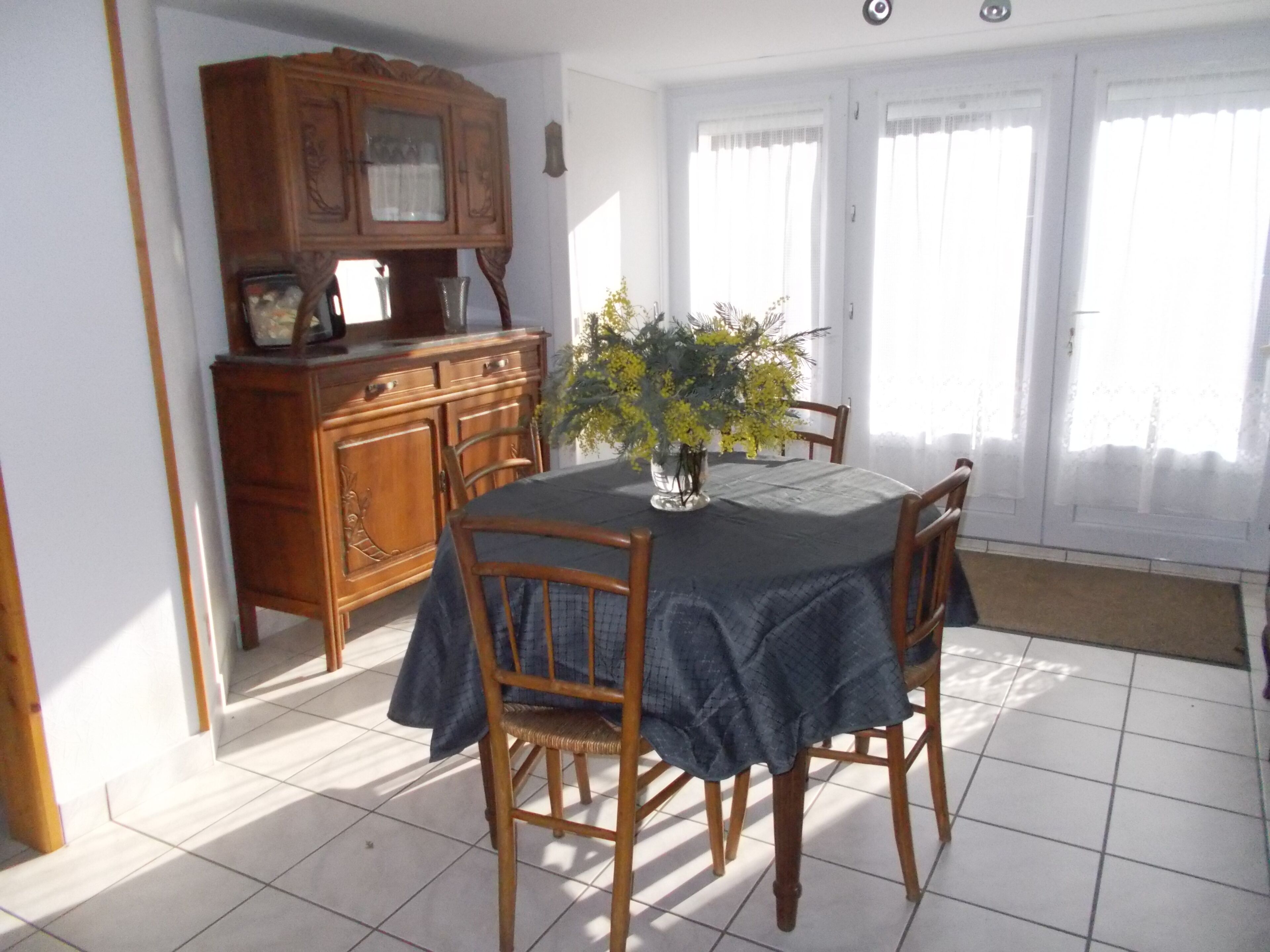 A dining area with a round table covered by a dark tablecloth, surrounded by wooden chairs. A vase with yellow flowers is placed on the table.
