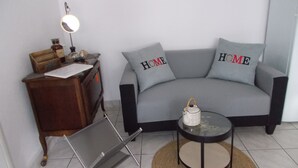 A living room with a grey sofa, two pillows with 'HOME' text, a small round table with a teapot, a wooden side table with a lamp, and a metal folding chair. - Charming, cosy villa 5 minutes' walk from the beach (Jullouville)