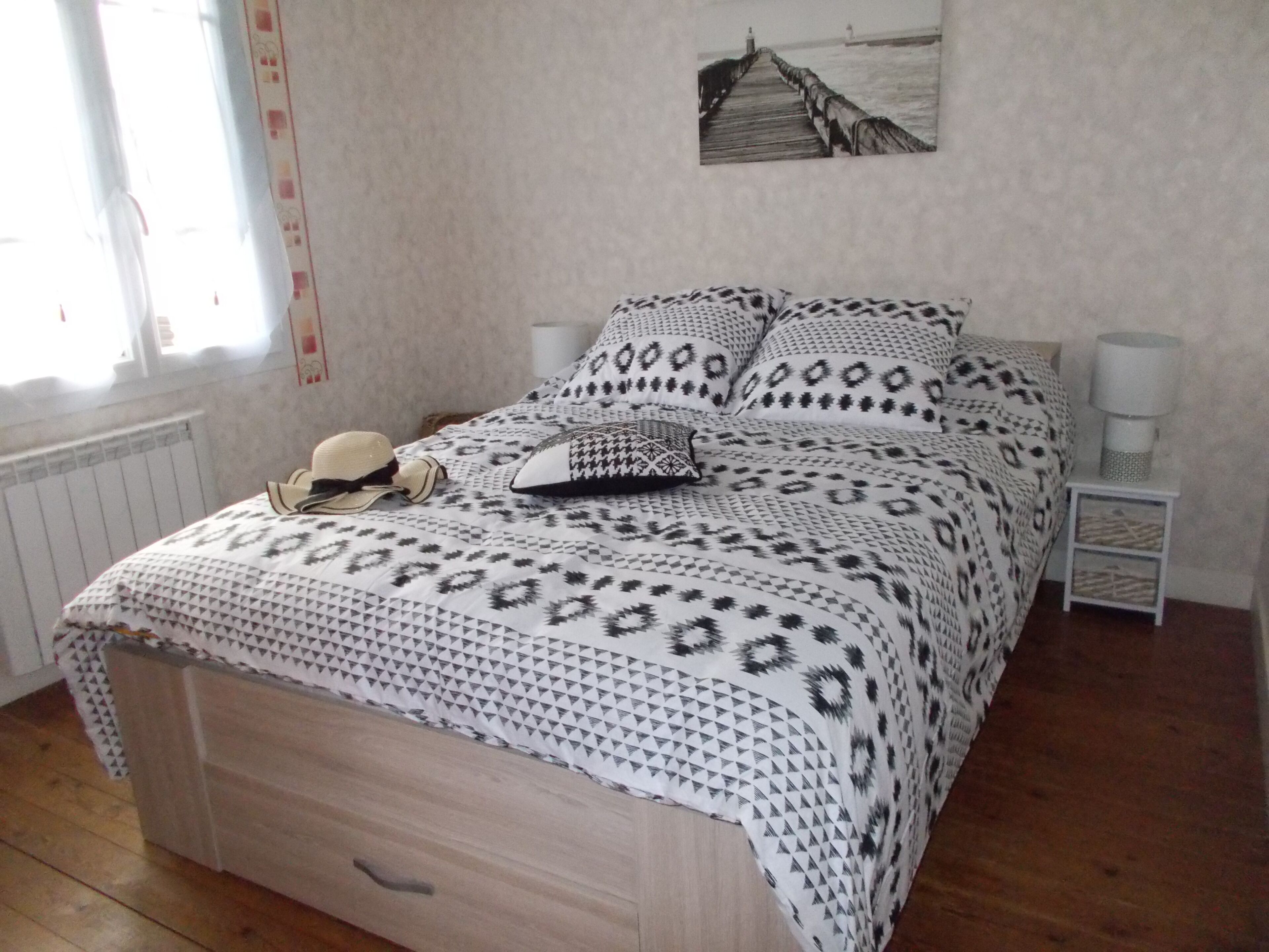 A bed with patterned bedding, a nightstand with a lamp, a wall-mounted picture, and a window with curtains.