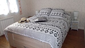 A bed with patterned bedding, a nightstand with a lamp, a wall-mounted picture, and a window with curtains. - Charming, cosy villa 5 minutes' walk from the beach (Jullouville)
