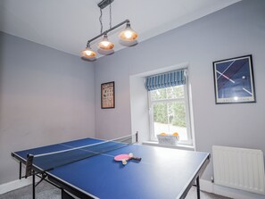 Una mesa de ping pong con red, paleta y pelota, una ventana con vistas, y un cuadro en la pared.