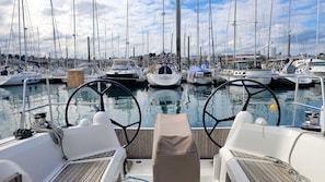 A marina with multiple sailboats docked, a clear sky, and a wooden deck with a steering wheel.