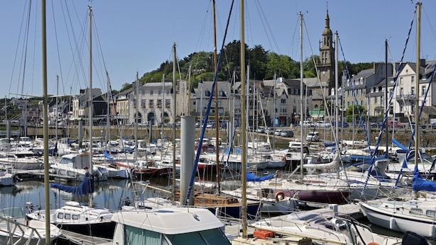 A marina with numerous boats docked, a prominent building with a tower, and a hillside with greenery in the background.