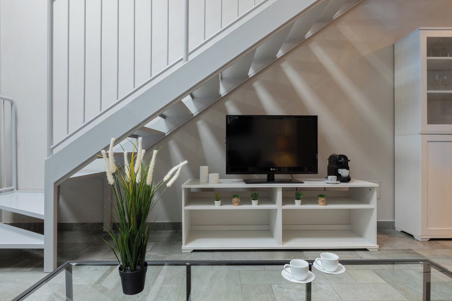 A modern living room with a staircase, a flat-screen TV, a coffee machine, and a glass table with cups.