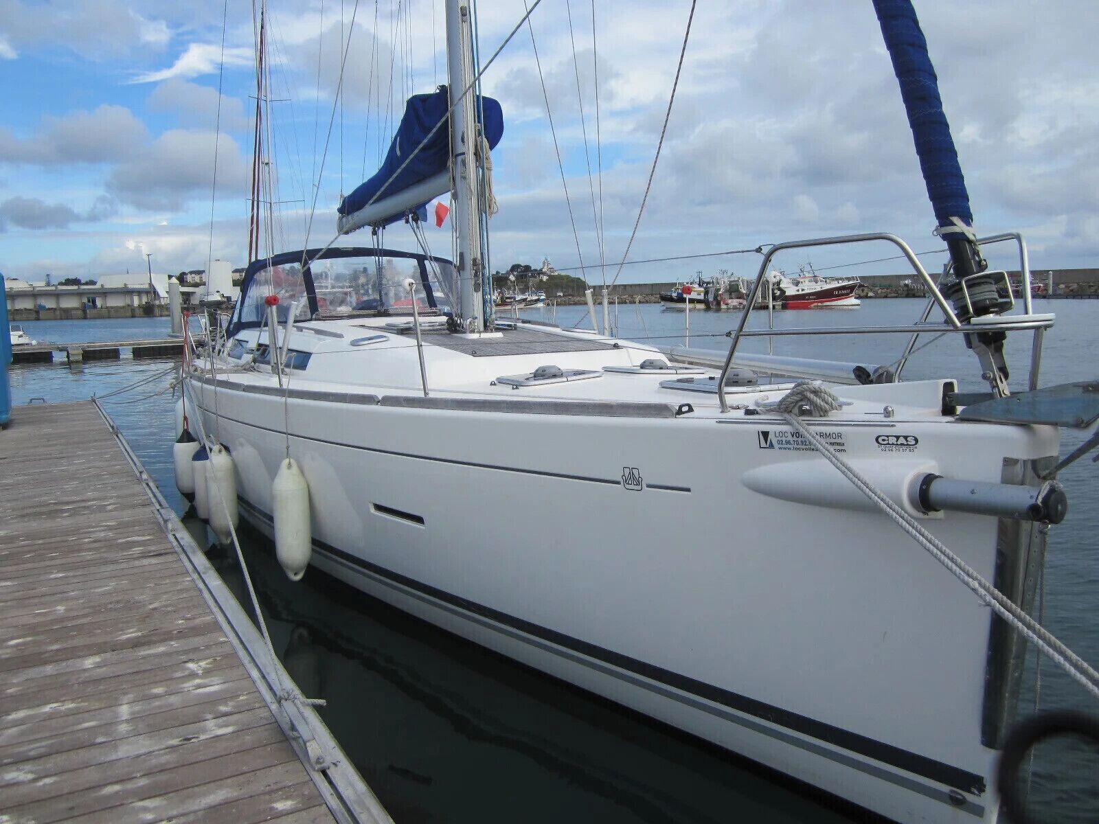 Ein Segelboot liegt an einem hölzernen Steg in einer Marina, der Himmel ist wolkenlos und im Hintergrund sind weitere Boote zu sehen.