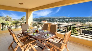 A balcony with a wooden table set for four, chairs, and a view of a town and hills. - Althea Villa | Private Pool, 5 Bedrooms & Garden Retreat Near Sandy Beach (Episkopi)