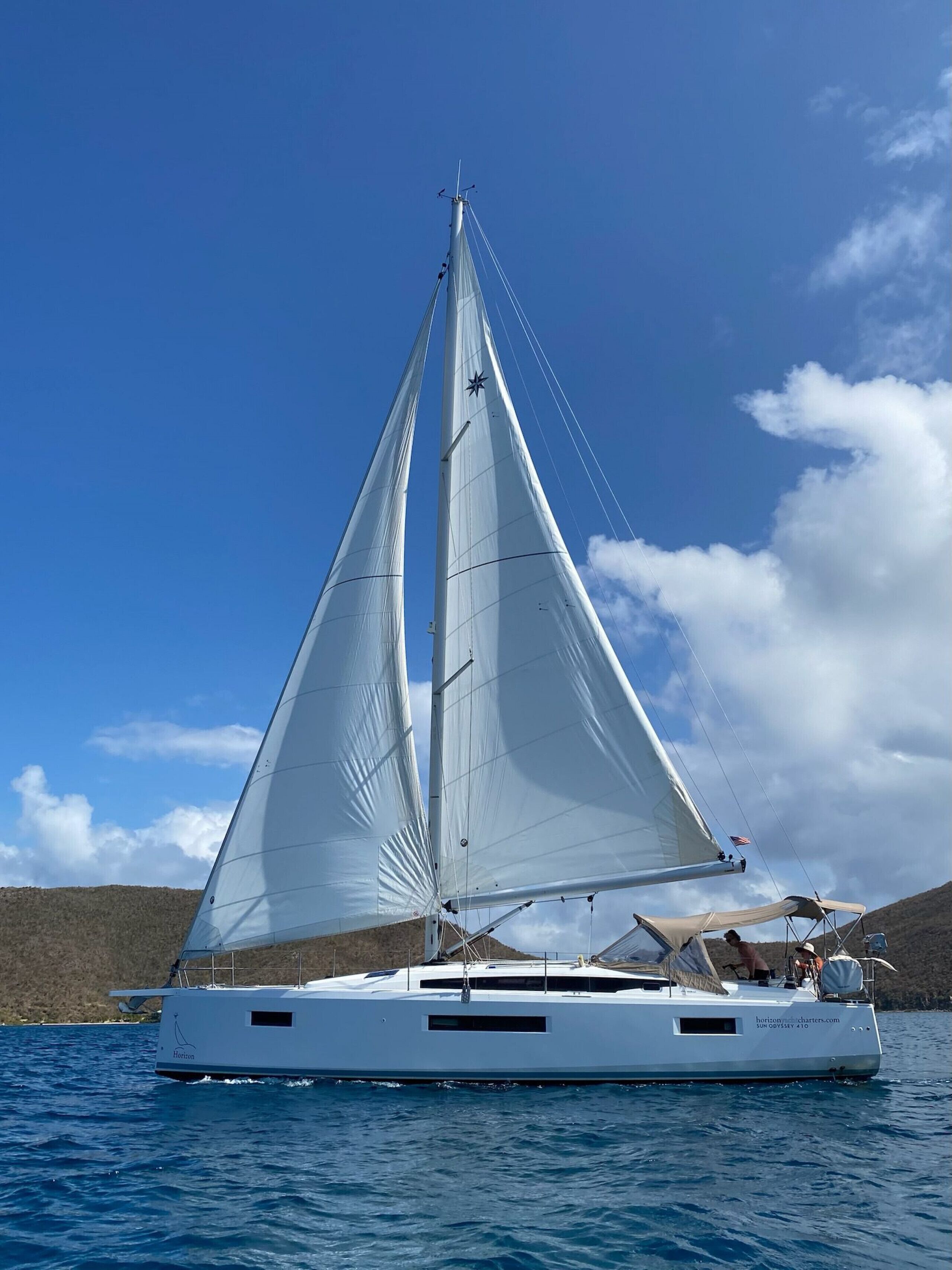 Un voilier à coque blanche et doté de nombreuses voiles, naviguant sur une étendue d’eau sous un ciel dégagé, avec des collines qui se dessinent à l’horizon.