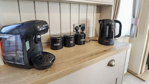 Une cuisine équipée d’une machine à café, de pots à sucre et à thé, et d’une bouilloire, le tout posé sur un plan de travail en bois.