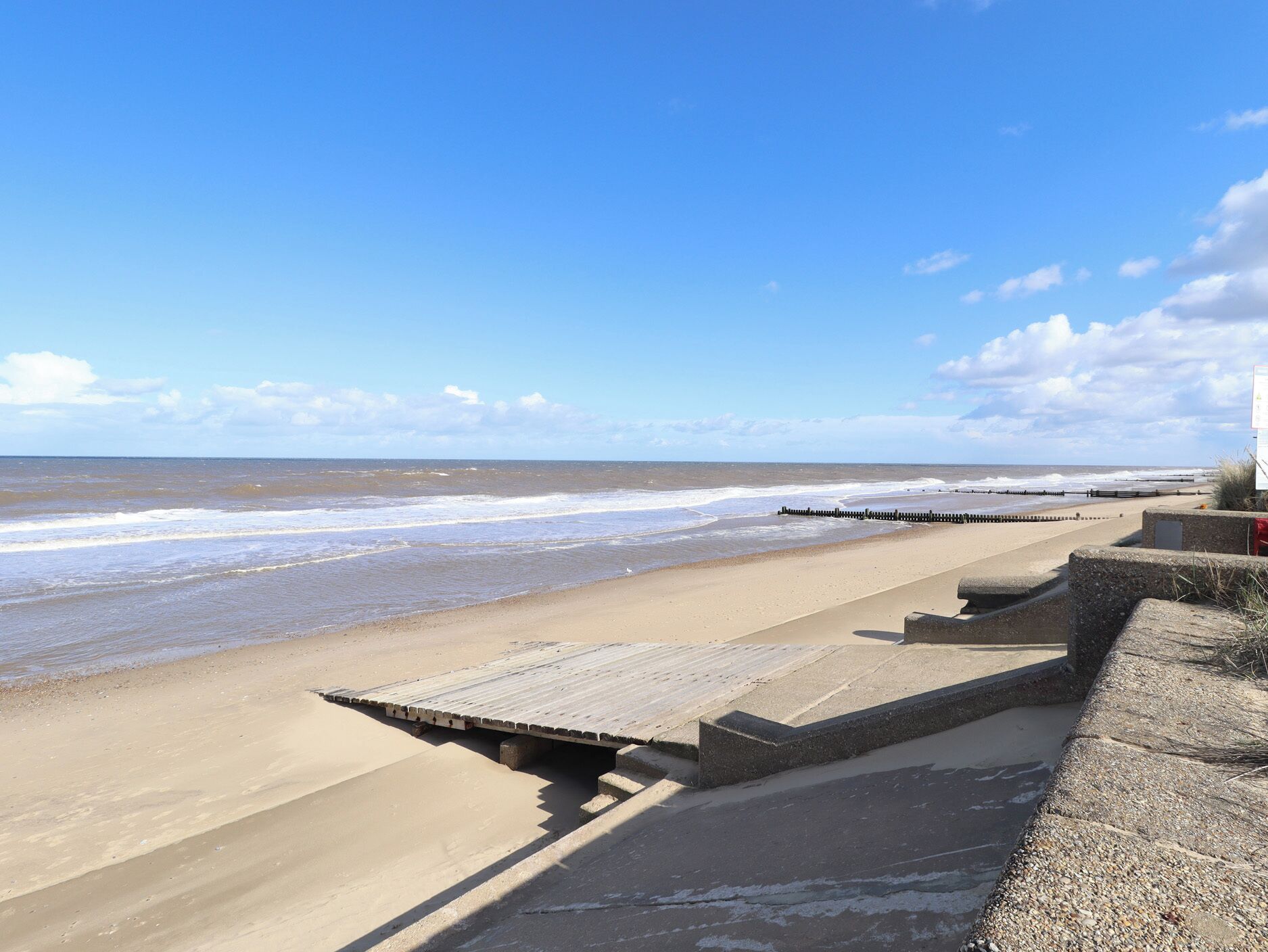 A beach with a wooden walkway leading to the water, a concrete barrier, and a clear sky.