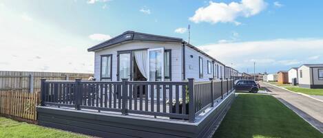 A mobile home with a fenced deck, a car parked in front, and a clear sky.