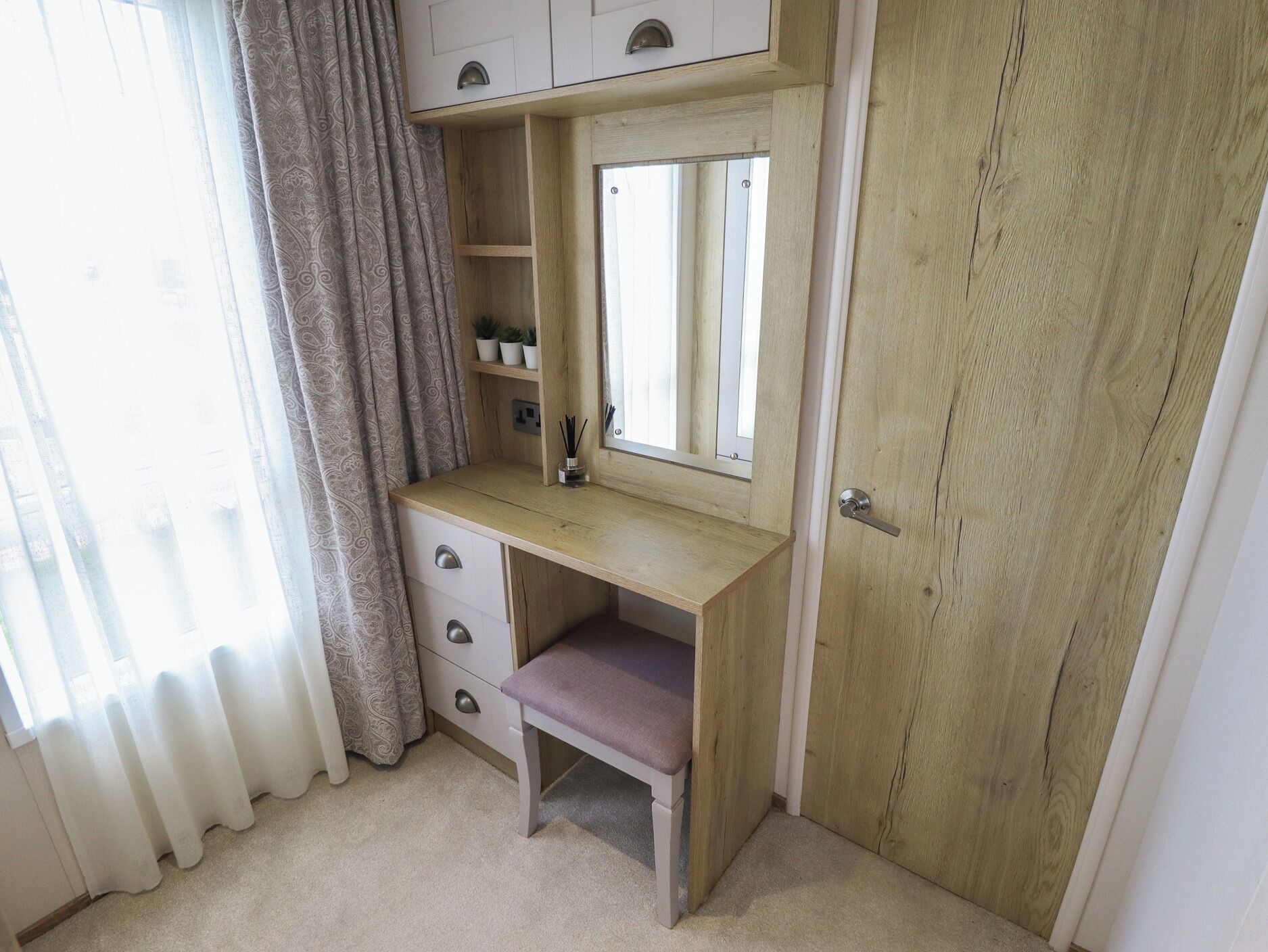 Un tocador de madera con espejo, repisa y cajón, en una habitación con puerta y cortinas.