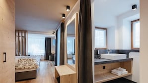 Una habitación de hotel moderna con piso de madera, cama con ropa de cama estampada, un espejo grande, un baño con lavamanos y bañera, y vista a la ciudad.