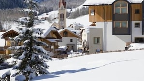 Un pueblo en la montaña cubierto de nieve con una iglesia destacada, casas de madera y un edificio moderno con grandes ventanales.