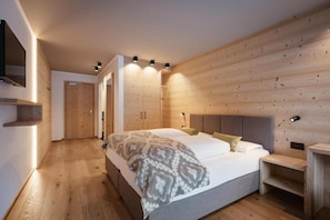 A modern bedroom with a wooden wall panel, a bed with pillows, a bedside table, and a TV mounted on the wall. - Berghotel Sanvi (San Martino in Badia)