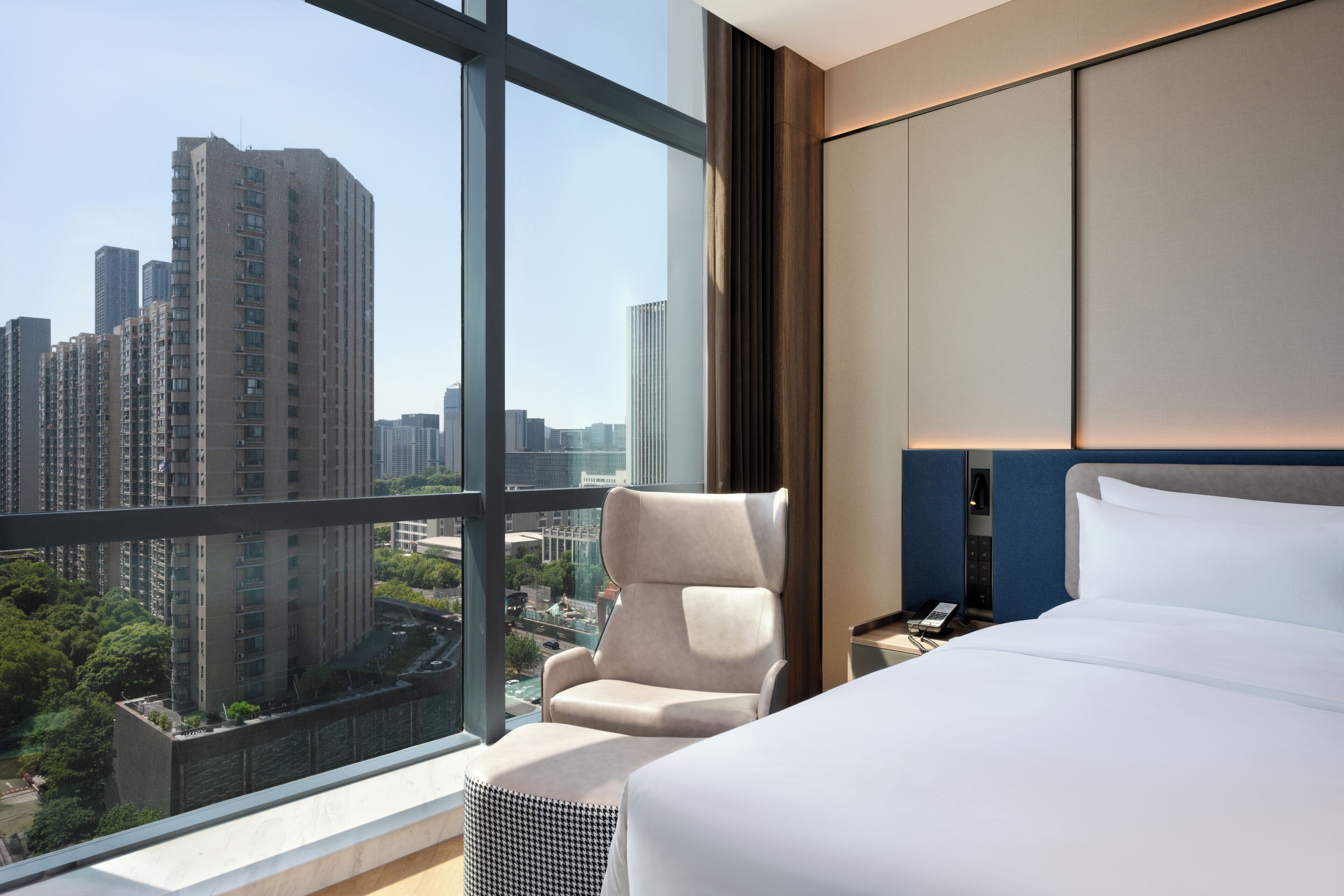 A hotel room with a large window, a bed, a chair, and a bedside table.
