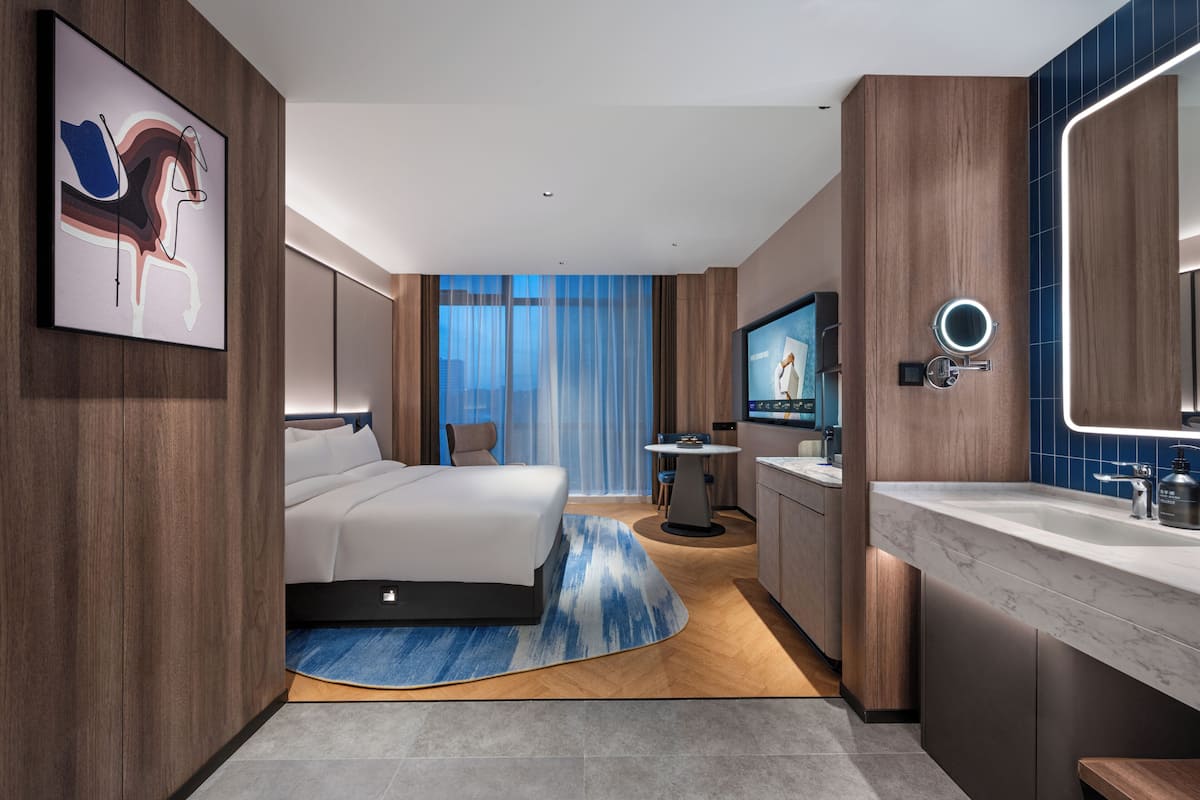 A modern hotel room with a bed, a desk, a round mirror, and a bathroom with a marble countertop.