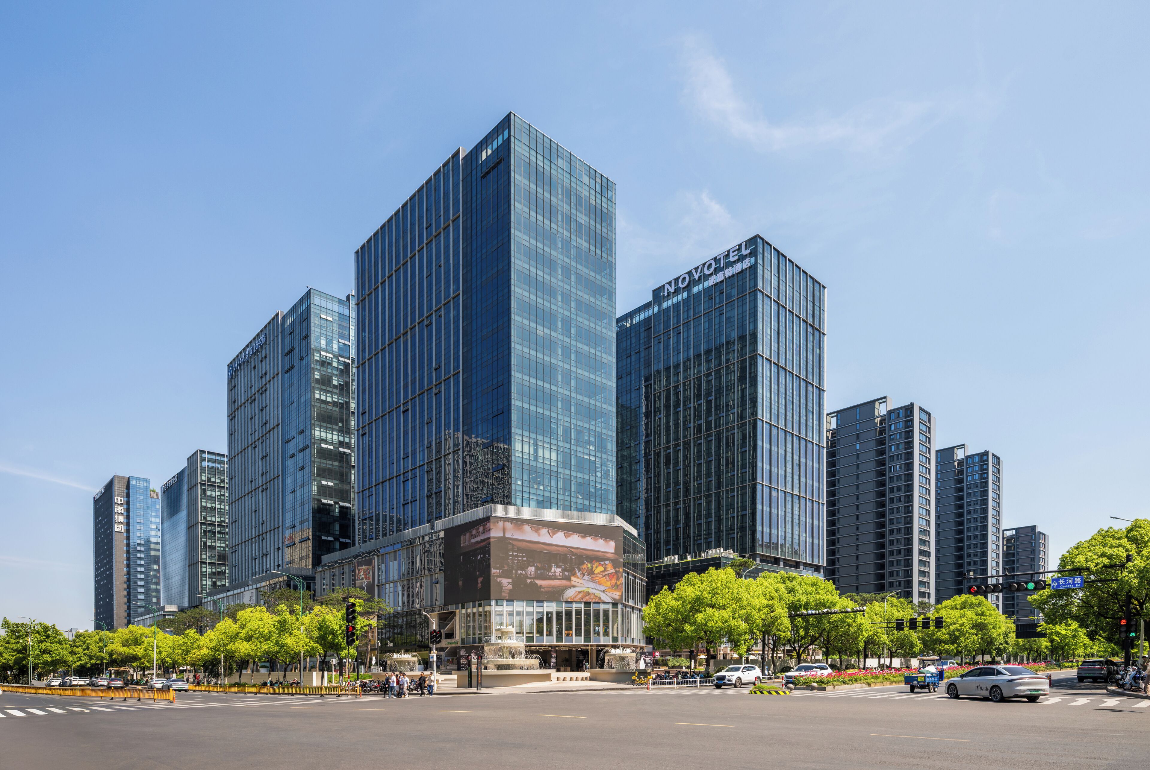 A modern hotel with a glass facade, located at a city intersection with traffic lights and a crosswalk.